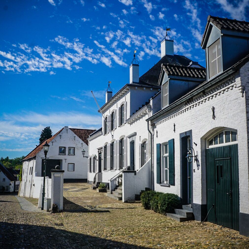 Een straat met kinderkopjes en witte huizen met donkere luiken onder een helderblauwe lucht met wolken. De huizen hebben traditionele Nederlandse architectuur met puntgevels en schoorstenen.