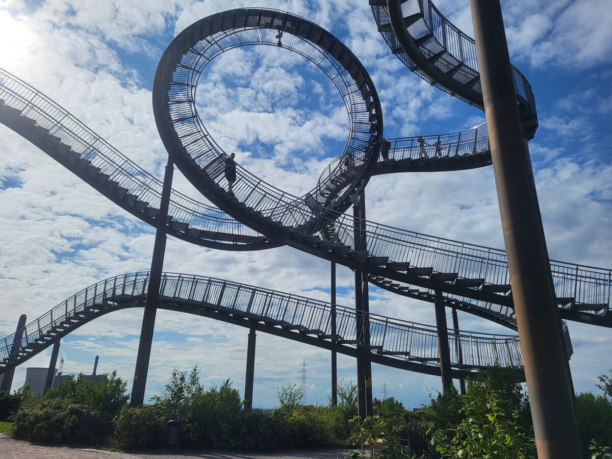 Een grote, stalen constructie met trappen en loopbruggen die in een spiraal en lusvorm omhooggaan tegen een blauwe lucht met witte wolken. Er zijn enkele mensen zichtbaar op de constructie. Op de achtergrond zijn bomen en struiken te zien, met verder weg industriële gebouwen en hoogspanningsmasten.