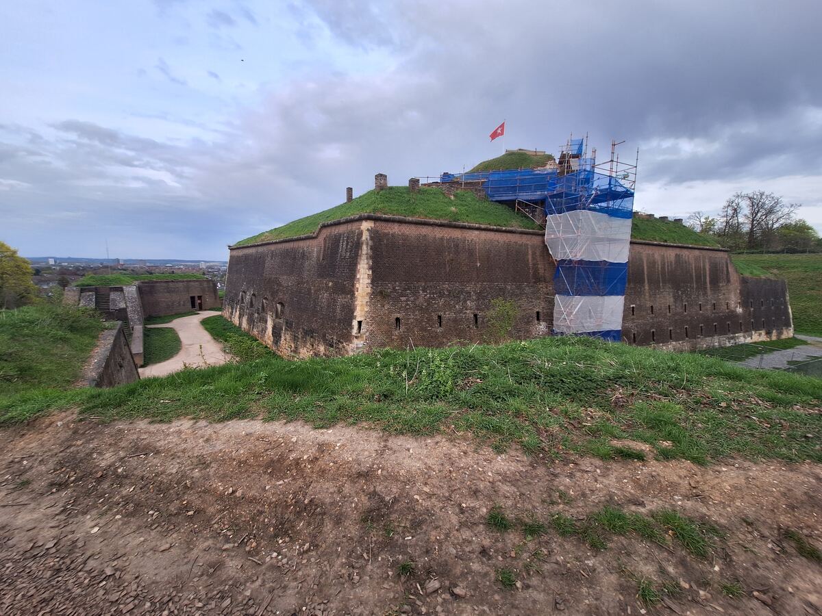 Een vestingwerk met bakstenen muren en een met gras begroeide bovenkant, deels in de steigers met blauwe en witte netten. Op de top van het fort wappert een rode vlag met een wit kruis. Op de achtergrond is een stad te zien onder een bewolkte lucht.