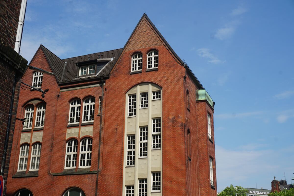 Een close-up van een groot, roodbakstenen gebouw met een complex dak en meerdere ramen. Het gebouw heeft diverse architectonische details, waaronder trapgevels, boogvormige ramen en een verticale strook van lichtere, mogelijk stenen, panelen met ramen in het midden. Op een hoek van het gebouw is een koperen element te zien met lichte oxidatie. De lucht is helderblauw met enkele kleine, witte wolkjes. Aan de linkerzijde is een deel van een ander bakstenen gebouw zichtbaar en rechtsonder wat groen