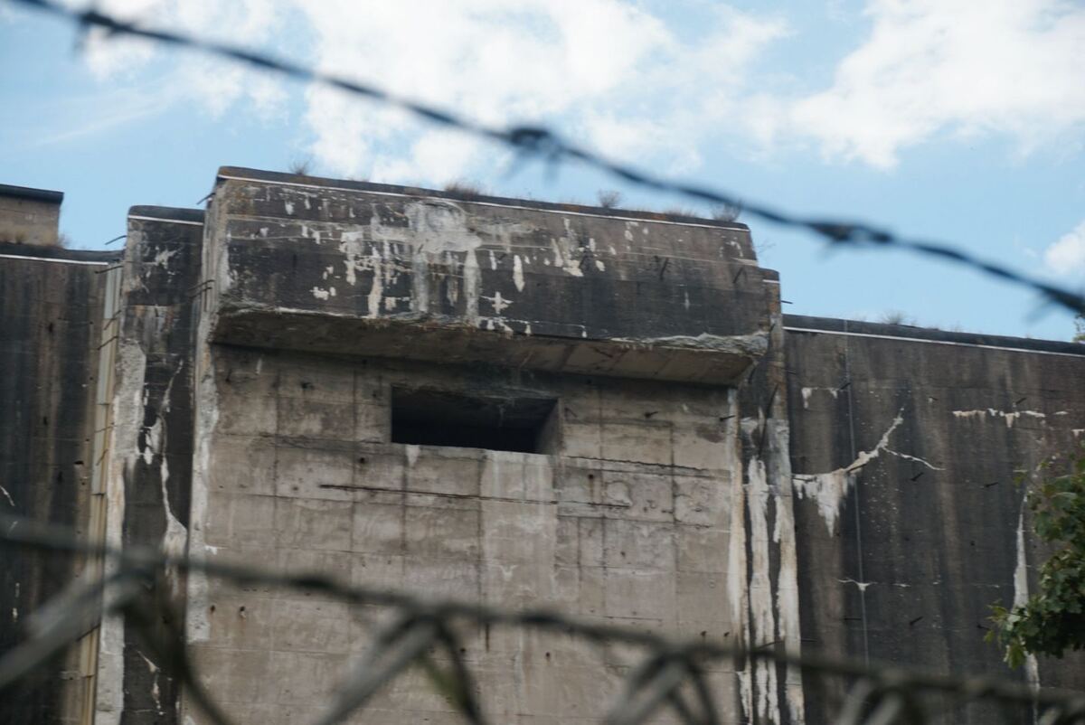 Een close-up van de massieve betonnen muren van Bunker Valentin, met een vierkante opening en zichtbare wapening. De bunker is deels begroeid met planten en er is prikkeldraad op de voorgrond te zien. De lucht is blauw met enkele wolken.