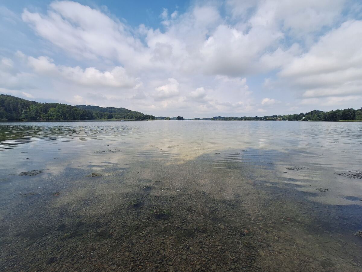 Een breed panorama van een kalm meer met een kiezelbodem die door het heldere water zichtbaar is. Aan de horizon zijn beboste heuvels te zien onder een helderblauwe lucht met verspreide witte wolken.