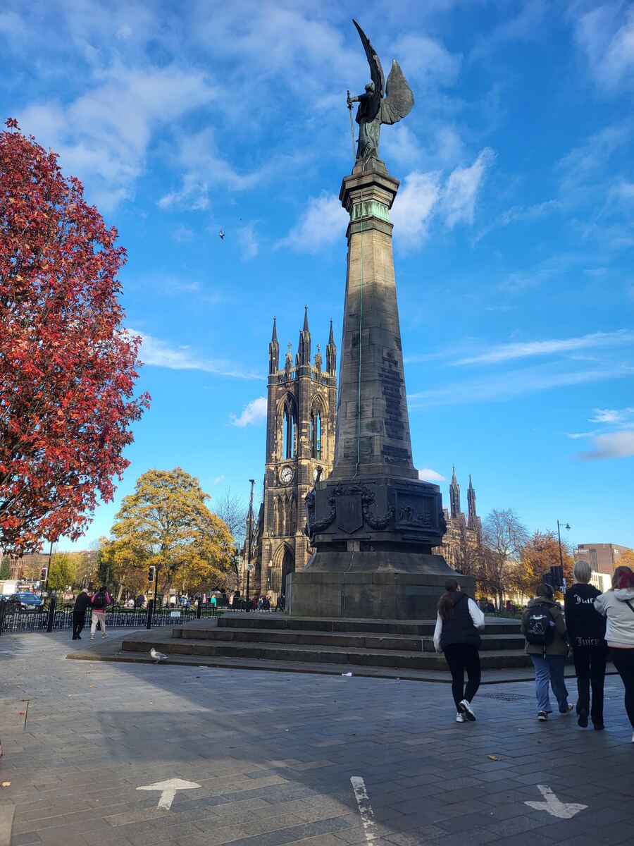 Een standbeeld van een engel met vleugels op een hoge sokkel staat centraal, met op de achtergrond een grote kerk met een klok en spitse torens. Links is een boom met rode herfstbladeren te zien en rechts lopen enkele mensen over een geplaveid plein. De lucht is blauw met witte wolken.