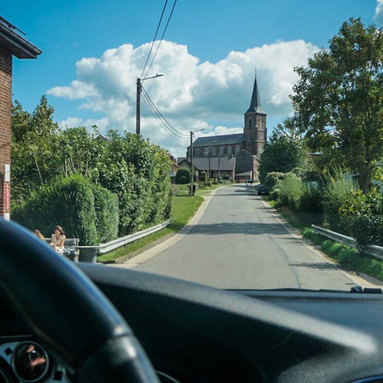 Een foto vanuit een auto die een smalle dorpsstraat inrijdt, met een kerk met een spitse toren op de achtergrond. Aan de linkerkant van de weg zit een persoon op een bankje. De lucht is blauw met witte wolken.