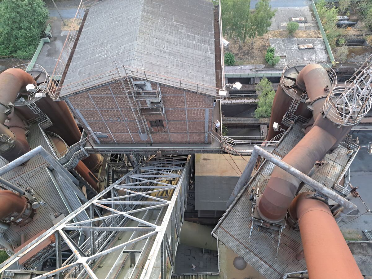 De foto toont een bovenaanzicht van een uitgestrekt industrieel complex met grote roestbruine pijpen, metalen constructies en bakstenen gebouwen. Er zijn ook betonnen oppervlakken en hier en daar groeit vegetatie tussen de structuren. Een persoon is zichtbaar op een van de loopbruggen.