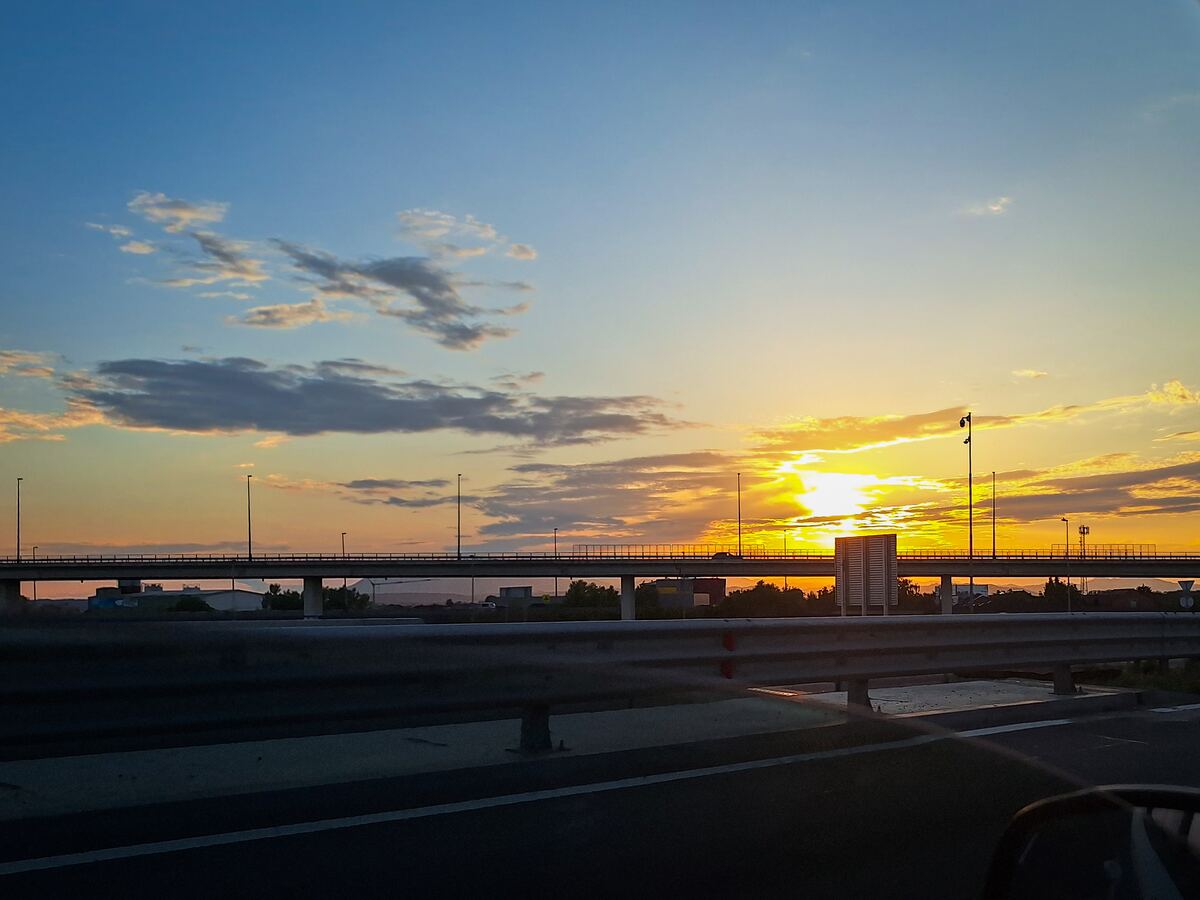De foto toont een zonsondergang boven een snelweg met een viaduct. De zon staat laag aan de horizon en kleurt de lucht oranje en geel, met blauwe tinten hoger in de lucht en enkele wolken. Op het viaduct zijn lantaarnpalen zichtbaar en op de snelweg op de voorgrond is een vangrail te zien.