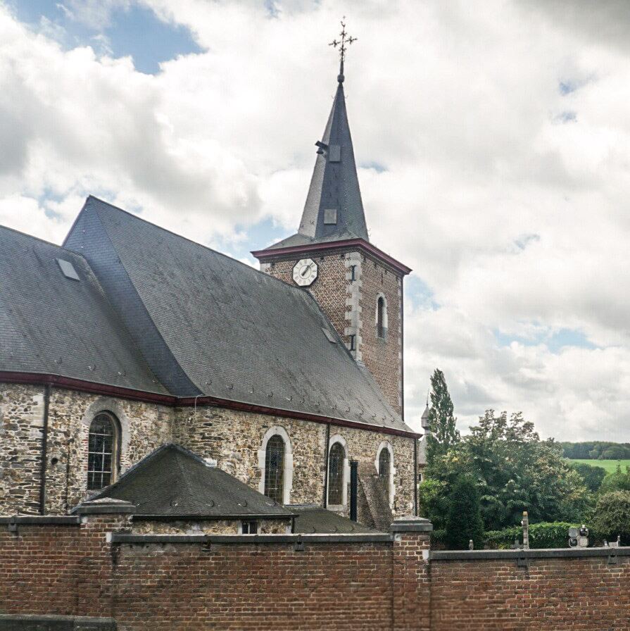 Een stenen kerk met een hoge, spitse toren en een klok aan de zijkant, gezien vanaf de zijkant achter een bakstenen muur. De kerk heeft een donkergrijs leien dak en ronde boogvensters. Op de achtergrond is groen landschap en een bewolkte lucht zichtbaar.