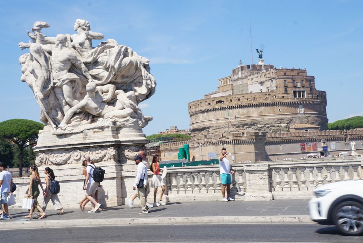 De foto toont het Castel Sant'Angelo in Rome, gezien vanaf een brug. Op de voorgrond staat een grote, witte marmeren sculptuur met meerdere figuren. Mensen lopen over de brug en op de stoep langs de weg. Rechtsonder is een deel van een witte auto zichtbaar. De lucht is helderblauw en de zon schijnt fel.