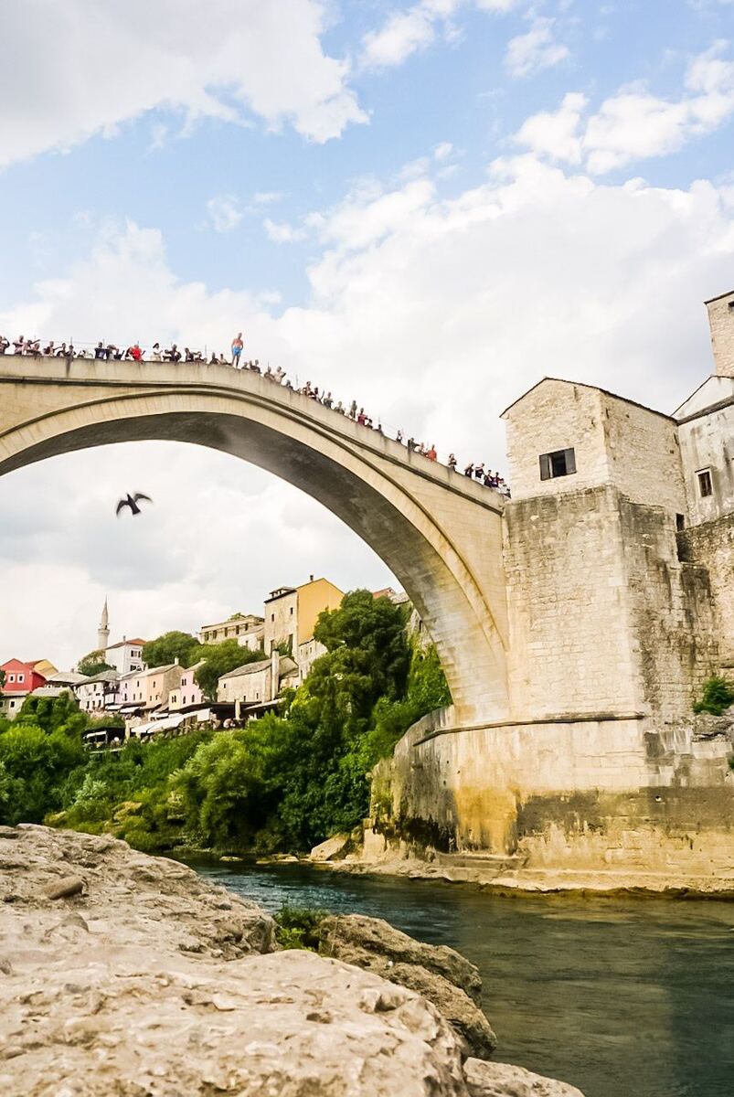 Een foto van de Stari Most brug in Mostar, met een man die zich klaarmaakt om te springen terwijl toeschouwers toekijken. Onder de brug stroomt een rivier en op de achtergrond zijn huizen en een minaret te zien onder een bewolkte lucht.