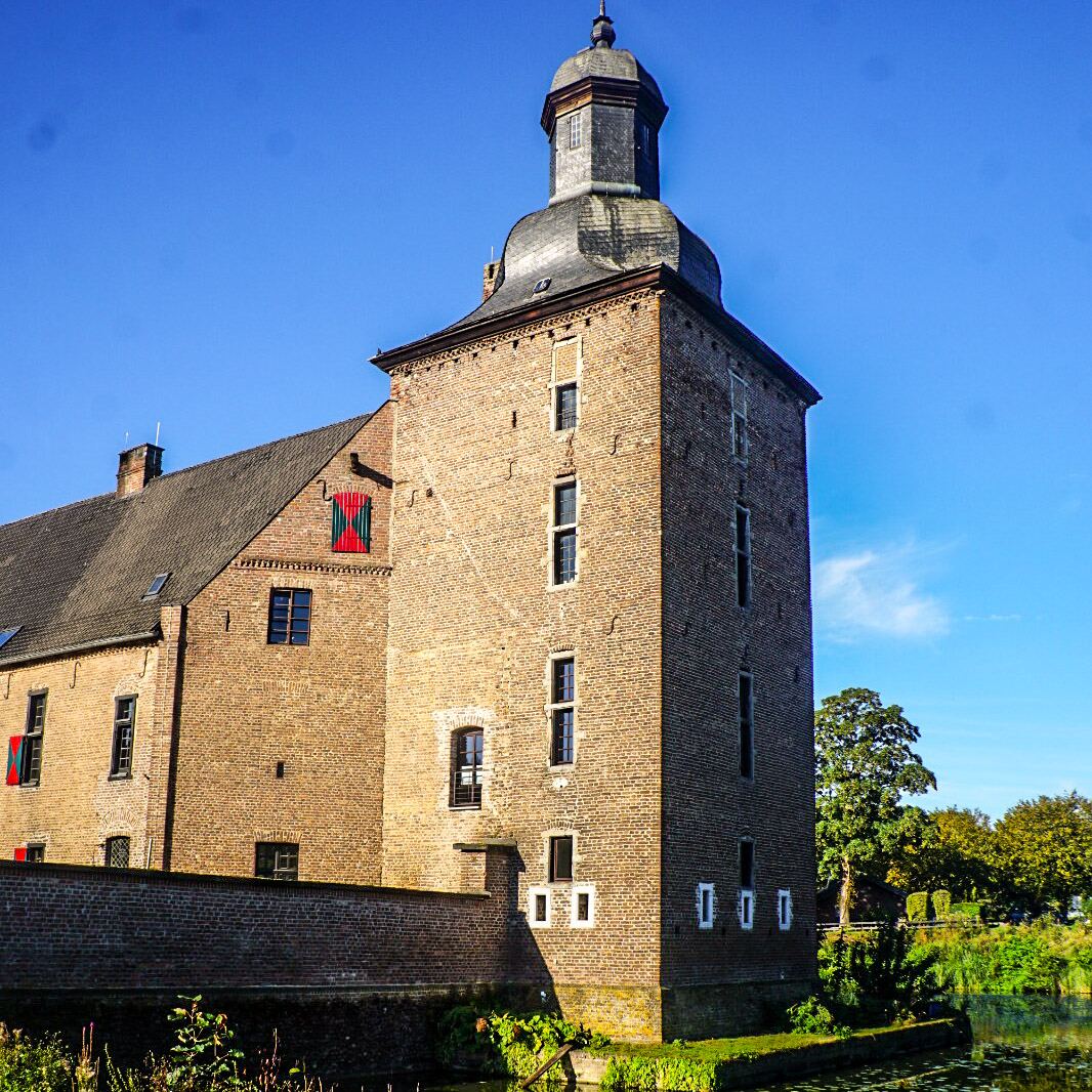 Een bakstenen kasteel met een toren en een gracht op een zonnige dag. De toren heeft een donker, achthoekig dak en er zijn ramen zichtbaar in de muren. Aan de linkerkant van de toren is een ander deel van het kasteel te zien met een schuin dak en ramen met rood-groene luiken. Een bakstenen muur loopt langs de gracht, die deels bedekt is met waterlelies. Op de achtergrond zijn bomen en een helderblauwe lucht te zien.
