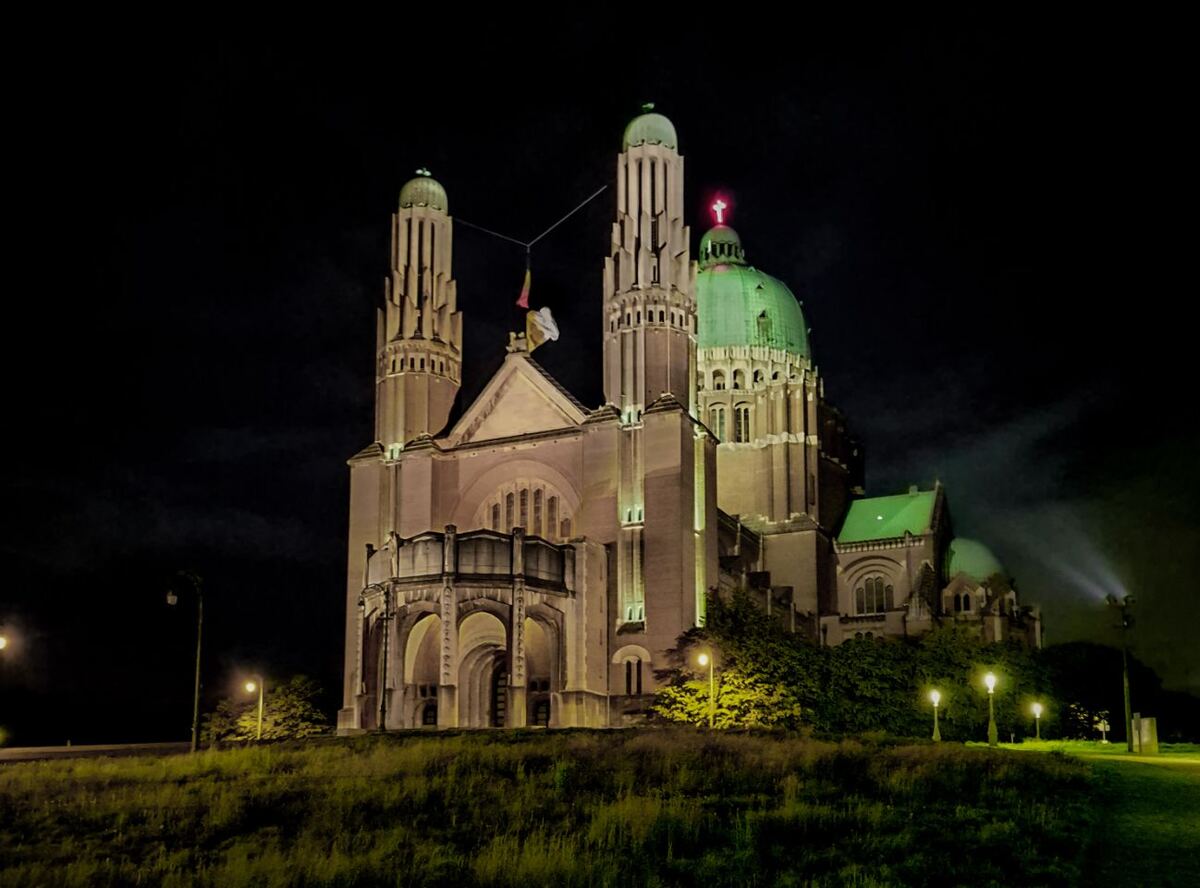 Een nachtopname van de Basiliek van het Heilig Hart in Koekelberg, Brussel, met de kenmerkende groene koepel en torens verlicht tegen een donkere hemel. De basiliek staat op een verhoging met gras en enkele bomen op de voorgrond, en er zijn straatlantaarns zichtbaar.