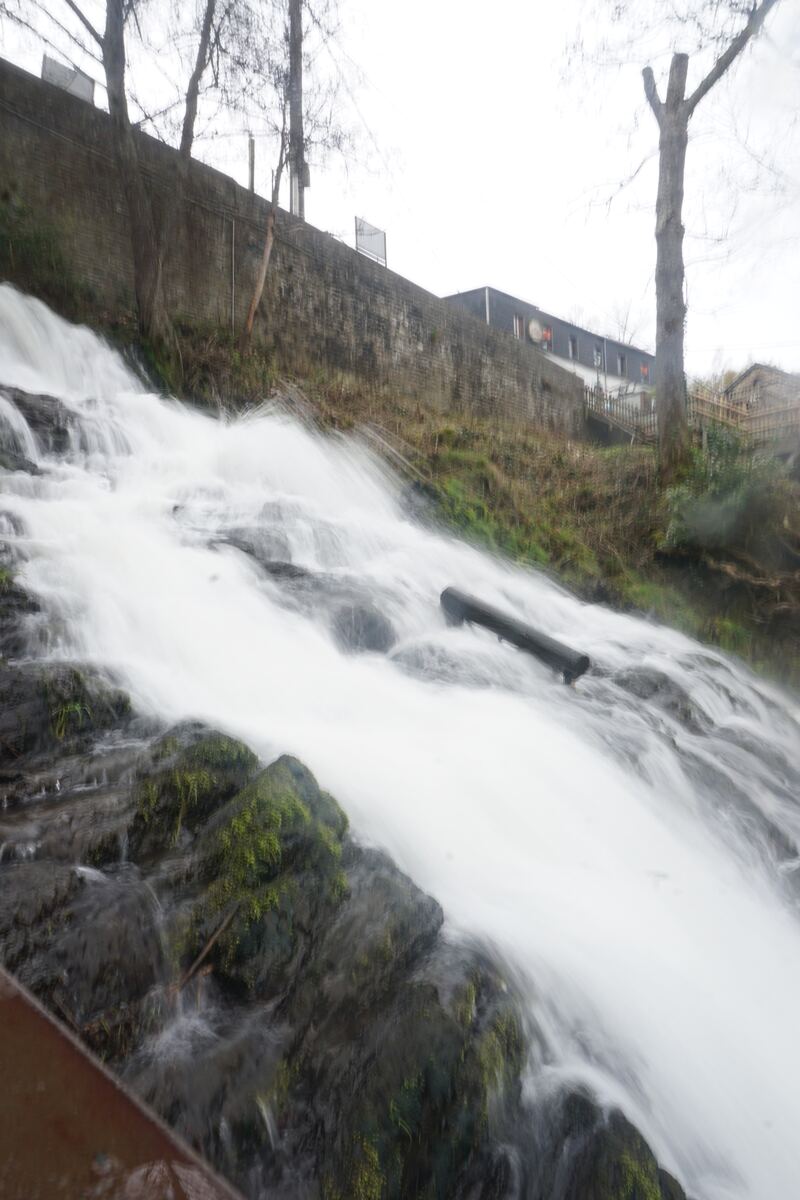 Een waterval stroomt over rotsen die bedekt zijn met mos, met op de achtergrond een stenen muur, kale bomen en een gebouw. Het water is lang belicht, wat een zacht, vloeiend effect geeft.
