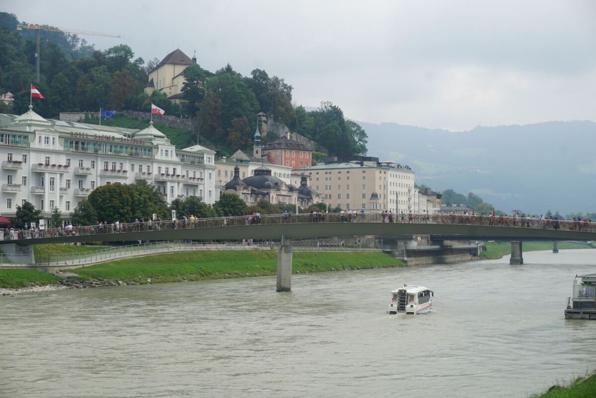 Een brede rivier met een bootje in het midden, een brug vol mensen en gebouwen langs de oever, waaronder 'Hotel Sacher Salzburg' met Oostenrijkse vlaggen, en heuvels op de achtergrond onder een bewolkte lucht.
