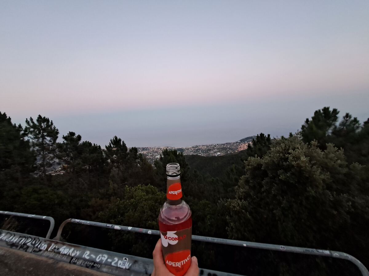 Een hand houdt een open fles 'Aperitivo' vast, met op de achtergrond een panoramisch uitzicht over een verlichte kustplaats en de zee. De voorgrond toont een metalen reling met graffiti en donkere boomtoppen. De lucht is in de schemering, met zachte blauw-roze tinten boven de horizon. De stad beneden is verlicht met talloze lichtjes.