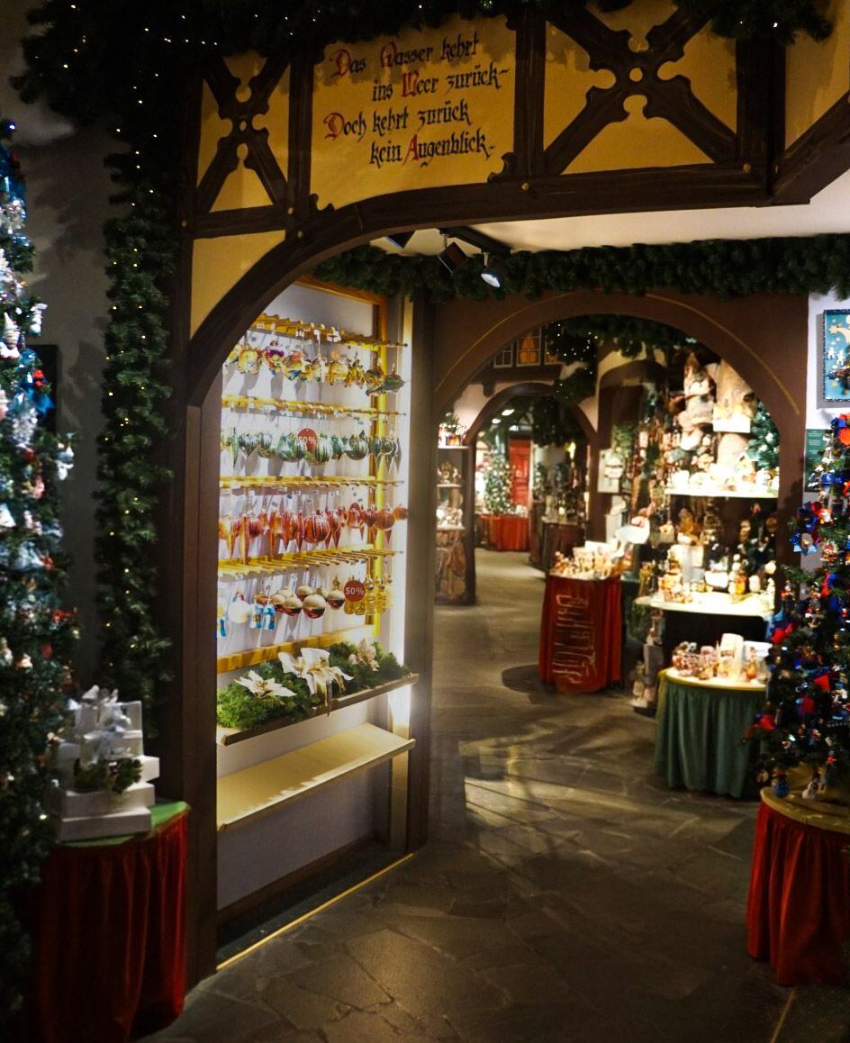 Een interieurfoto van een winkel met kerstversiering. Links staat een kerstboom met cadeautjes eronder. In het midden is een display te zien met diverse kerstballen en ornamenten. Boven de doorgang hangt een houten bord met Duitse tekst. Op de achtergrond zijn meer versierde kerstbomen en tafels met kerstdecoraties zichtbaar.