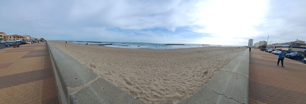 Een panoramische foto van een breed zandstrand met een boulevard en de zee. Aan de linkerkant staan gebouwen en geparkeerde auto's langs de boulevard. Rechts op de foto is een gebouw met de tekst 'CASINO' te zien. De lucht is deels bewolkt en de zee heeft lichte golven. Er zijn enkele personen op het strand en de boulevard.